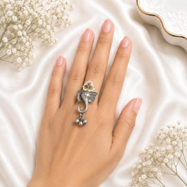 Hand wearing an oxidized German silver Ganesha statement ring featuring hanging little silver beads from Kreezo, displayed on a soft white background with floral accents.