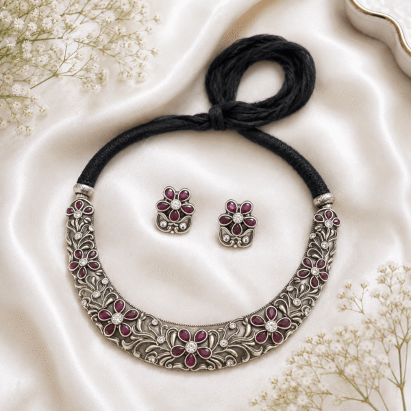 Kreezo oxidized silver rigid choker necklace and matching floral stud earrings featuring intricate filigree vine carvings, ruby-red stone petals, and sparkling white stone centers on a white satin background with baby's breath flowers