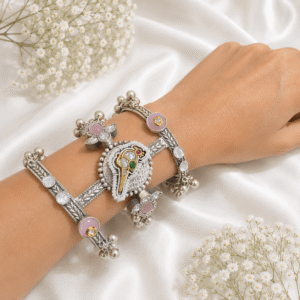 A single, wide, german silver-tone kada bracelet featuring an ornate central section with Kundan stone settings, green and red accents, pearls, and dangling metallic beads, resting on a white satin background.