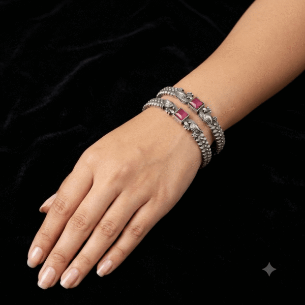 Model wearing a pair of Kreezo oxidized silver bangles detailed with intricate filigree and a prominent square ruby-red stone against a black velvet background