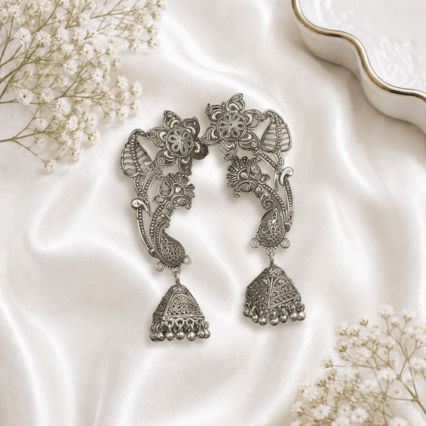 A pair of ornate oxidized silver jhumka earrings featuring intricate floral and paisley filigree work with hanging jhumka bells and ghungroos, beautifully arranged on a soft white satin fabric alongside delicate baby's breath flowers.
