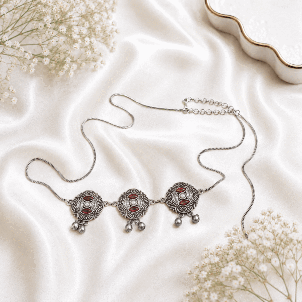 An intricate oxidized silver kamarbandh (waist chain) featuring three medallion motifs embedded with red and white stones and small dangling drops, beautifully displayed on a white satin fabric with delicate baby's breath flowers.