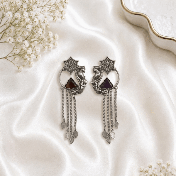 Pair of Kreezo oxidized silver statement earrings featuring an intricate star motif, perched peacock and circular frame surrounding a dark triangular stone, and multiple dangling chain strands with leaf charms displayed on a white satin background with baby's breath flowers