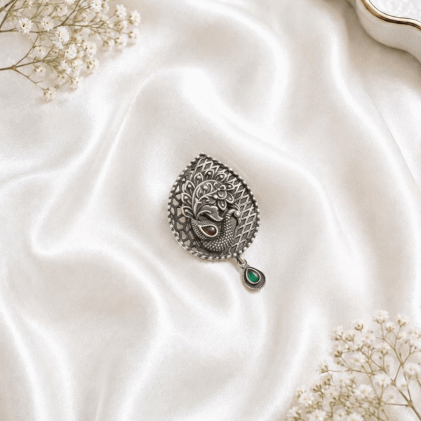 An intricate Peacock silver saree pin featuring a mandala flower design with red ruby-like stones, seed pearl detailing, and a hanging teardrop charm, displayed on a cream satin cloth with baby's breath flowers.