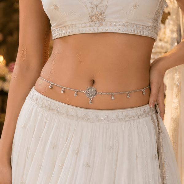 An intricate oxidized silver kamarbandh (waist chain) featuring three medallion motifs embedded with red and white stones and small dangling drops, beautifully displayed on a white satin fabric with delicate baby's breath flowers.