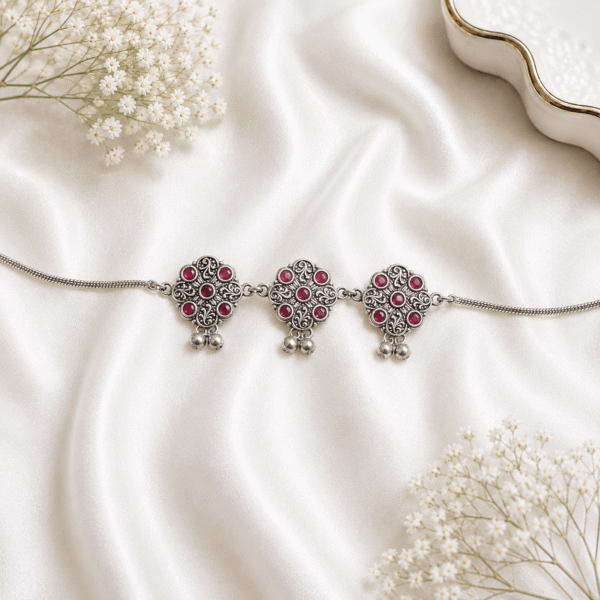 An intricate oxidized silver kamarbandh (waist chain) featuring three medallion motifs embedded with red and white stones and small dangling drops, beautifully displayed on a white satin fabric with delicate baby's breath flowers.