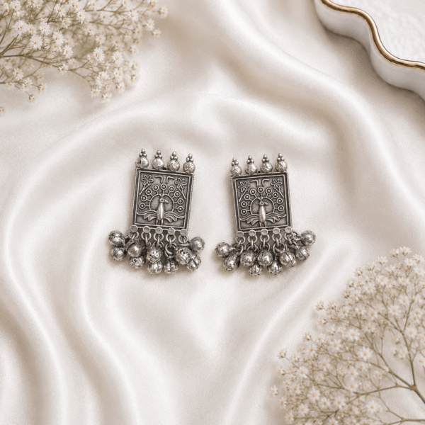 Pair of Kreezo oxidized silver rectangular stud earrings featuring an intricate carved peacock motif, floral accents, and a rich cluster of hanging silver ghungroo bells displayed on a white satin background with baby's breath flowers