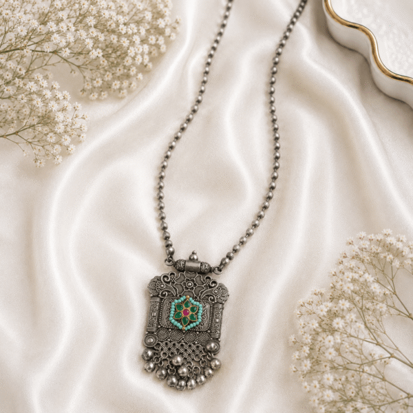 Kreezo oxidized silver beaded chain necklace with an intricate rectangular heritage pendant featuring filigree work, a central floral motif with ruby and green stones, turquoise bead details, and hanging silver ghungroo dangles on a white satin fabric with baby's breath flowers