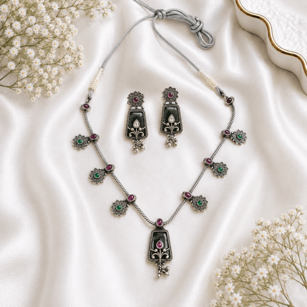 Kreezo silver necklace set featuring black enamel elements, floral motifs set with ruby-red and green stones, a central pendant, and matching drop earrings displayed on a white satin background with baby's breath flowers