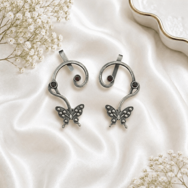 Pair of handcrafted antique oxidized silver ear cuffs featuring swirled vine bands, detailed butterfly bottom charms, and round dark red garnet stone studs, set against a soft white silk background.