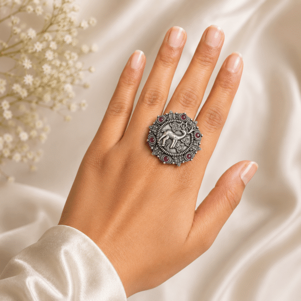 A hand wearing a round, silver ring with an engraved deer motif surrounded by small red ruby-like stones, resting on a cream satin cloth with baby's breath flowers.