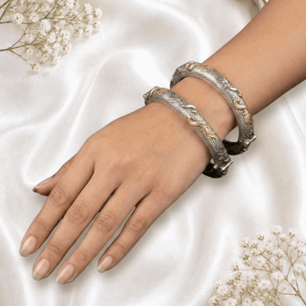 A wrist wearing a pair of textured silver and gold two-tone bangles featuring intricate golden leafy scrollwork, displayed on a soft cream satin cloth with baby's breath flowers.