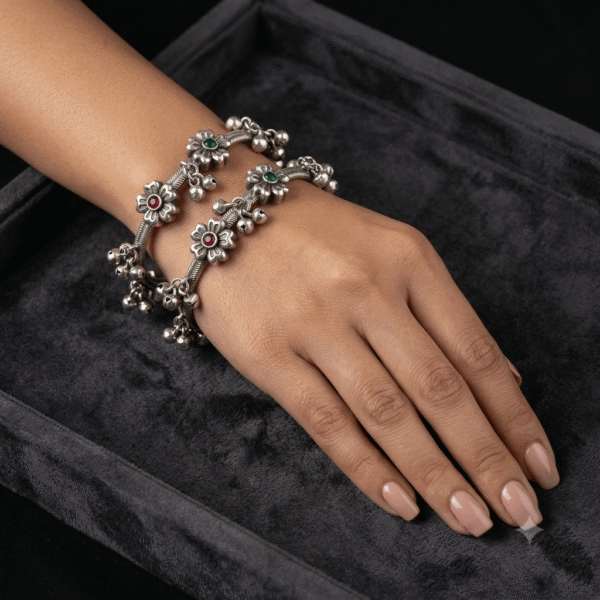 A pair of intricate silver bangles featuring floral motifs with dark stone centers and hanging ghungroo bells, held on a soft white fabric background.