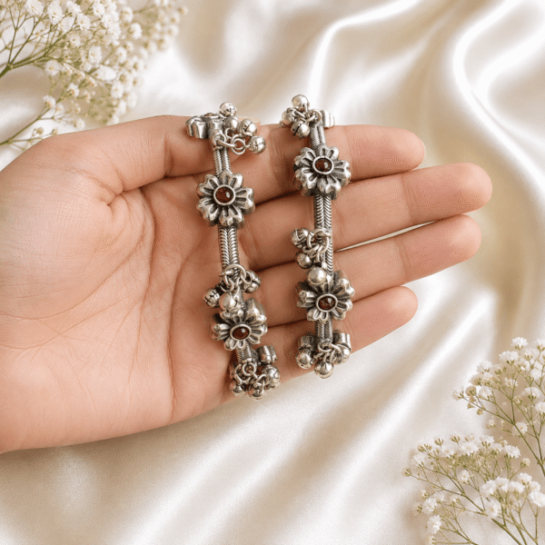 A pair of intricate oxidized silver bangles featuring floral motifs with dark stone centers and hanging ghungroo bells, held on a soft white fabric background.
