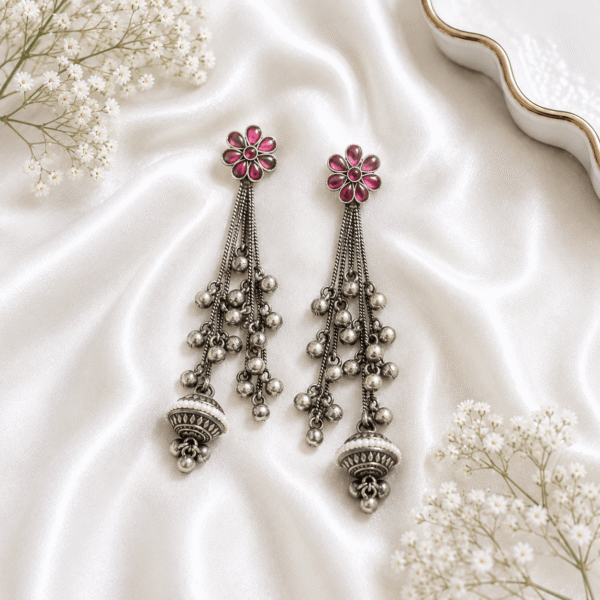 Pair of Kreezo oxidized silver long tasselled earrings featuring a floral stone stud with green and pink crystals and multiple hanging beaded chains displayed on a white satin background