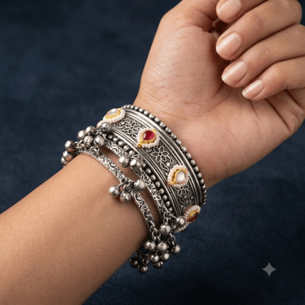 A wrist wearing a stacked set of intricate silver bangles featuring floral carvings, colored stone accents, and hanging ghungroo bells.