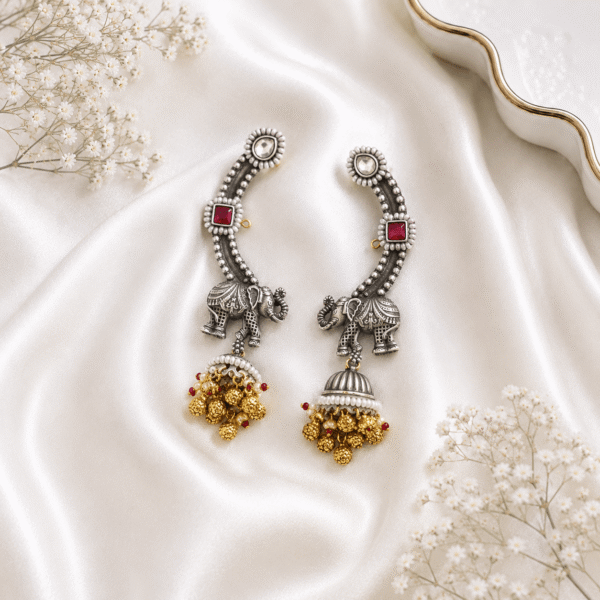 Pair of Kreezo oxidized silver statement ear cuffs featuring an intricate royal elephant motif, square ruby-red stones, white crystal studs, and dangling golden jhumkas displayed on a smooth white satin fabric with baby's breath flowers