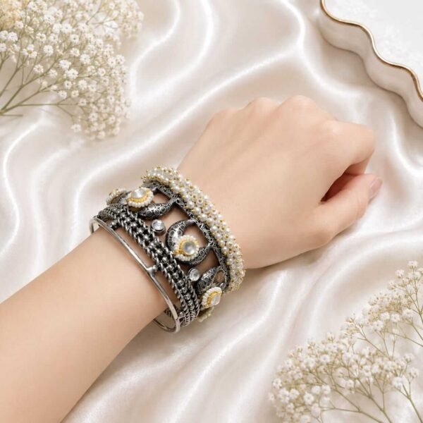A woman's wrist wearing an oxidized silver-tone intricate patterned kada paired with stacked pearl beaded bracelets on a white satin background.