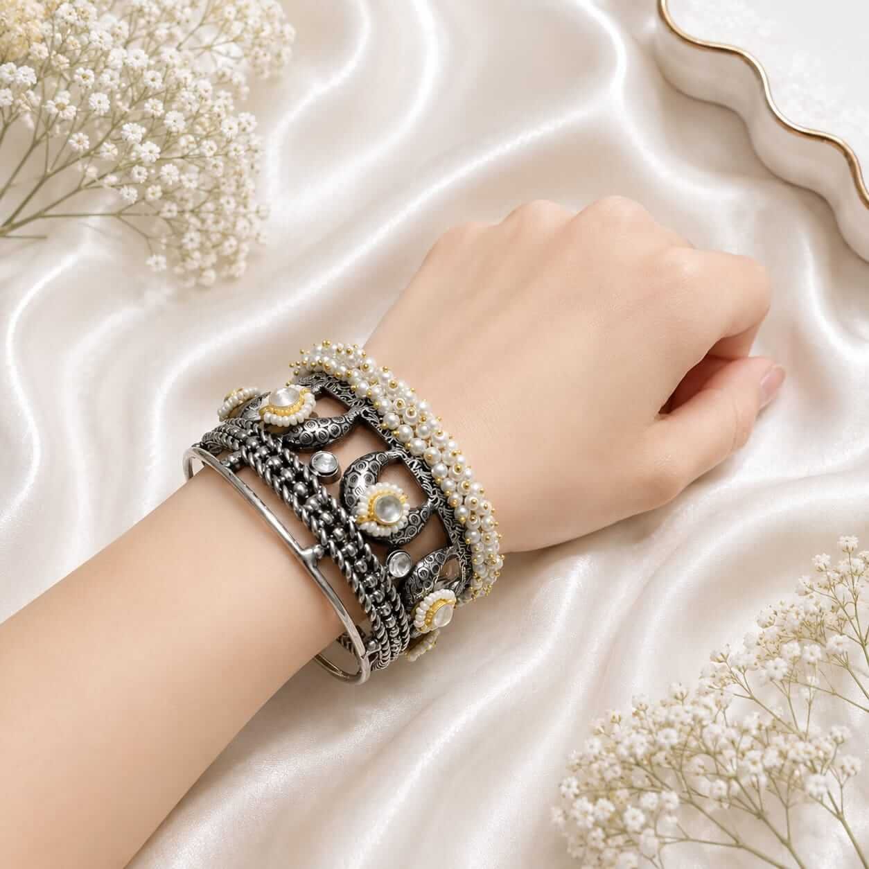 A woman's wrist wearing an oxidized silver-tone intricate patterned kada paired with stacked pearl beaded bracelets on a white satin background.