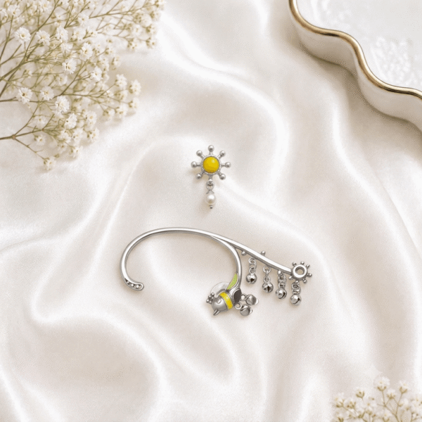 An artistic flat-lay shot of an oxidized silver jewelry set on a soft white satin fabric, featuring a sun-and-pearl stud earring and a matching bird motif ear cuff with dangling ghungroo chains, accompanied by delicate baby's breath flowers.