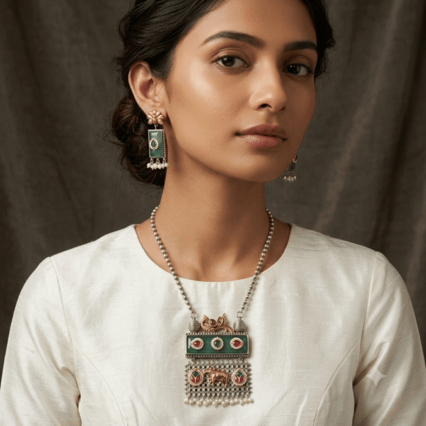 Model showcasing an artisan silver necklace and drop earrings set, featuring vibrant green rectangular stones, intricate multi-colored temple motifs, and white pearl bead fringe on a rich textured background.