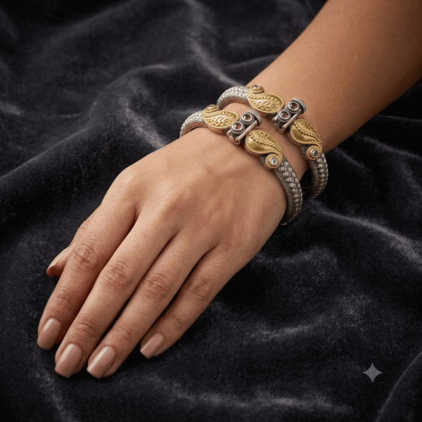 A wrist wearing a pair of textured silver and gold two-tone bangles featuring ornate mango (paisley) motifs studded with small ruby-like stones, displayed on a dark velvet background.
