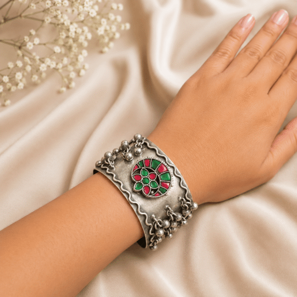 A wrist wearing a wide silver cuff bracelet featuring a circular motif with red and green meenakari stones, wavy borders, and hanging ghungroo bells, displayed on a soft satin fabric background.