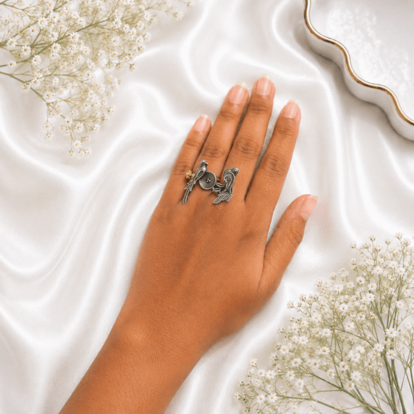 A hand wearing a unique oxidized silver adjustable ring featuring two facing bird motifs with detailed tail feathers and a central circular element, set against a black display background.