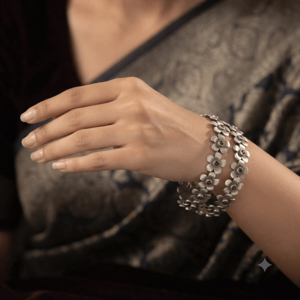 A person's hand holding a pair of oxidized silver link bangles designed with connected floral motifs, featuring dark red stones at the center of each flower against a dark background.