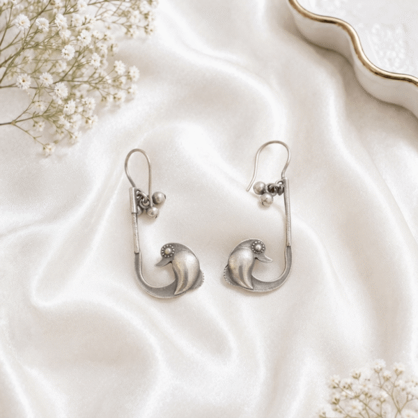 A pair of oxidized silver ear cuffs featuring a stylized duck motif at the base, dangling small bead charms near the hook, displayed on a soft white satin fabric background with baby's breath flowers.
