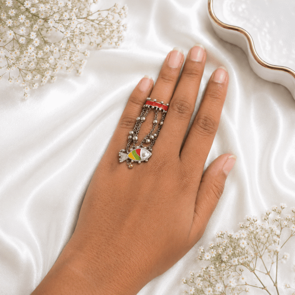 A hand wearing a unique oxidized silver hatha ring featuring a red enamel band connected by beaded chains to a colorful fish motif pendant, displayed against a white satin and baby's breath flower background