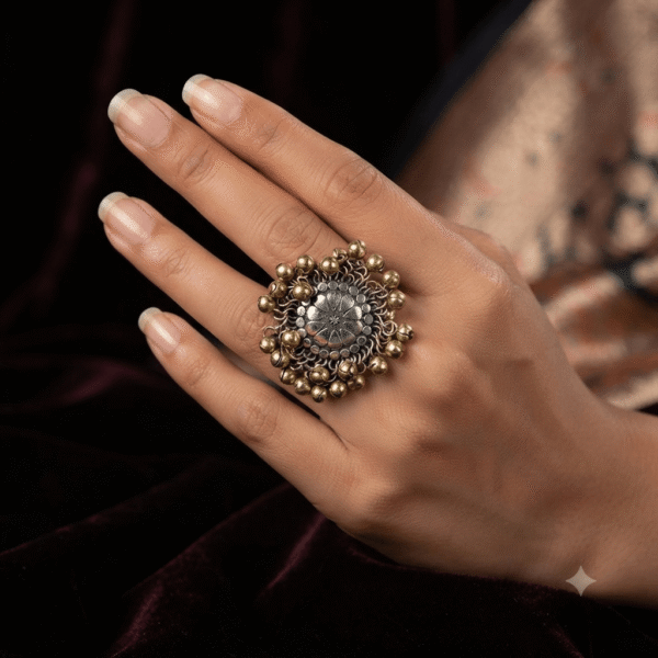 A hand wearing a large, round statement ring featuring an oxidized silver floral center surrounded by cascading gold-tone bead charms, against a dark velvet background.