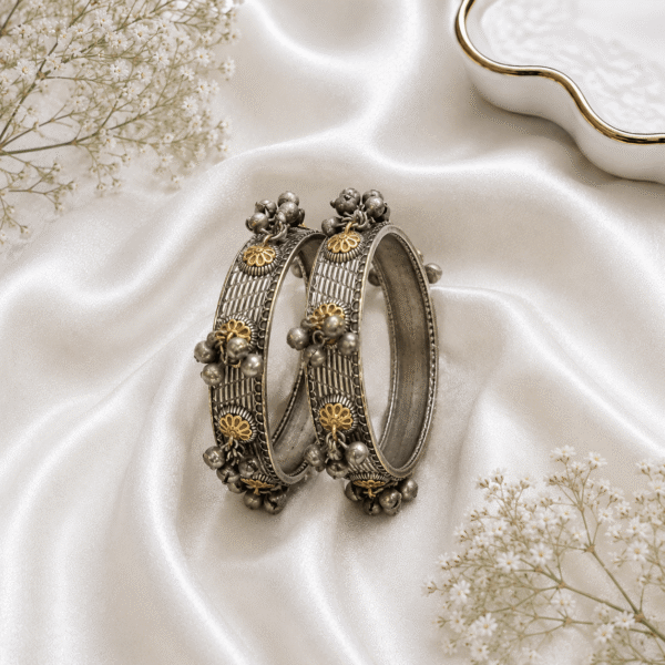 A pair of textured oxidized silver bangles featuring gold-tone floral motifs and cascading small ghungroo bell charms along the borders, placed on a white satin fabric background with white baby's breath flowers.
