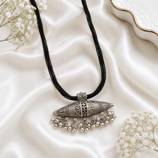 An intricate oxidized silver tribal-style pendant suspended from a multi-strand black cord necklace, featuring dangling small metal bead charms along the bottom, displayed on a soft white satin fabric background with white baby's breath flowers.