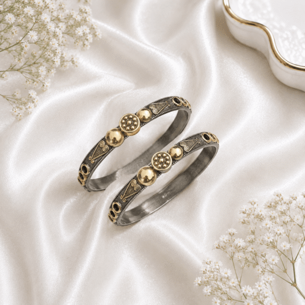 A pair of ethnic bangles featuring an oxidized silver base with intricate etched patterns and raised gold-tone bead and floral motifs, neatly arranged on a glossy white satin background with baby's breath flowers.